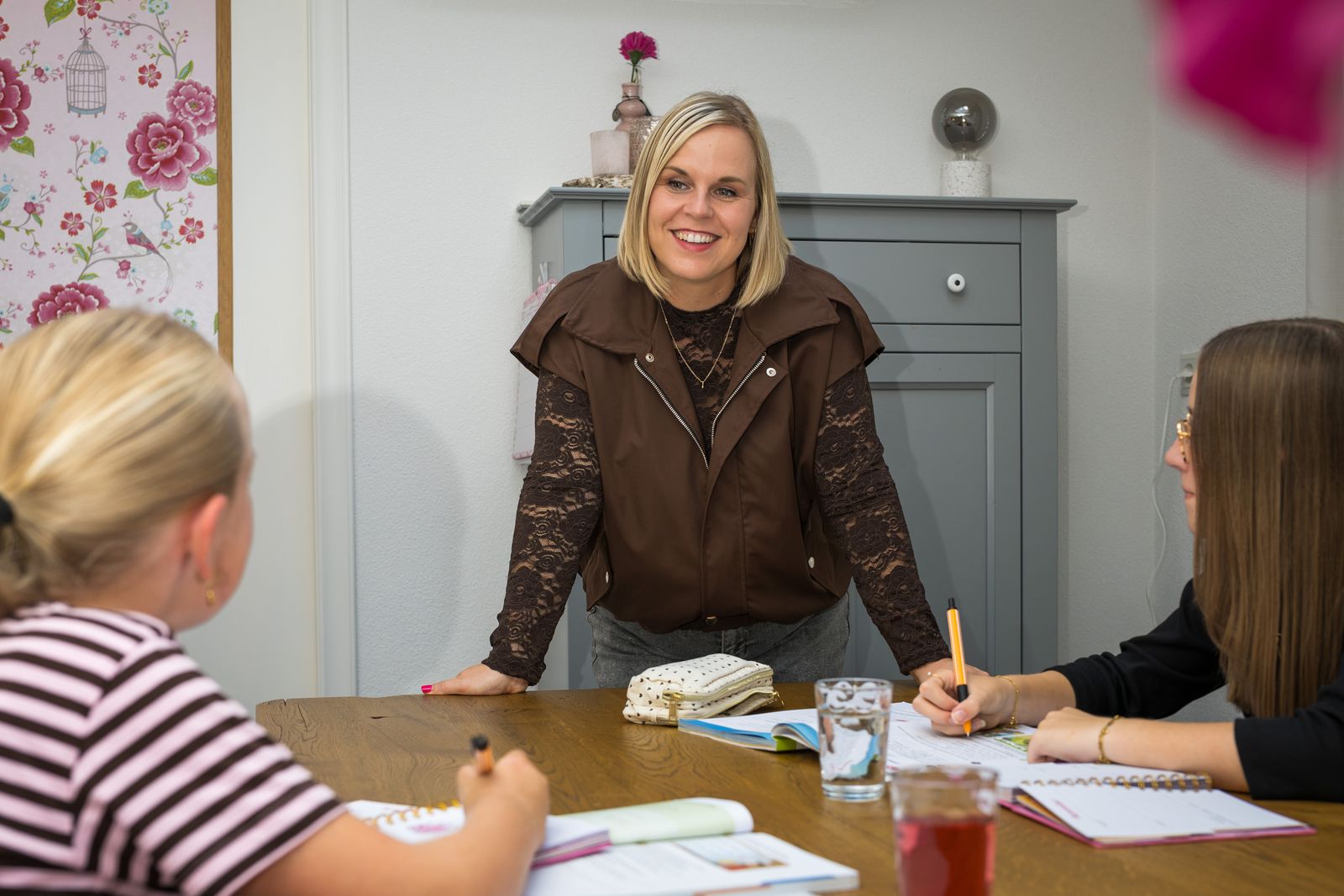 Suzanne staat lachend aan tafel, twee leerlingen werken in hun agenda en boeken